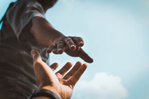 Two people reaching toward one another with outstretched hands against a bright sky, symbolizing support, connection, and meeting people where they are.